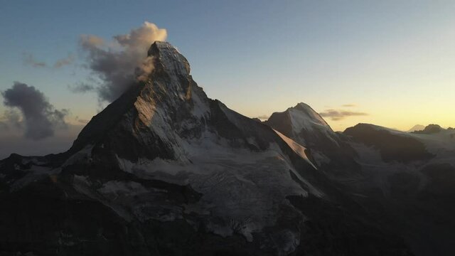 Drone shot moving forward with moving and dancing clouds changing form and shape as they stick and stay around the pointy snowy peak of the mountain Matterhorn in Switzerland during an epic sunrise.