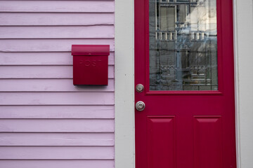 The entrance to a quaint country style cottage with a fuchsia colour antique decorative glass door, a vintage letterbox or postal box on a pink exterior clapboard wooden wall of a residence. 
