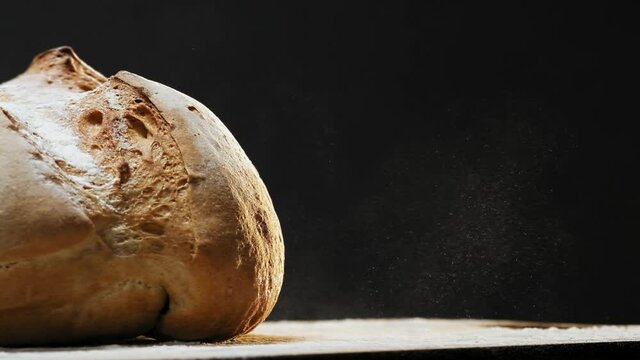 Loaf of homemade wheat bread with white flour falls down onto dark table on black background extreme close view slow motion