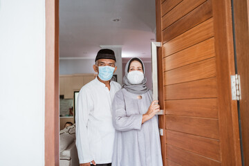 Fototapeta na wymiar muslim mature couple wearing face mask standing at front door of their house with greeting gesture