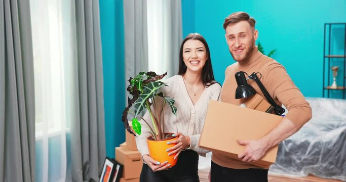 Portrait of cheerful young happy cute family man and woman move in holding cardboard box and palnt in hands. American couple chatting and discussing decor of house unpacking stuff. looking at camera