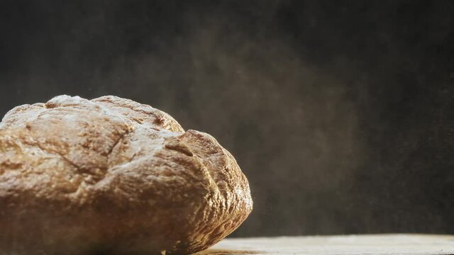 Tasty homemade rye bread loaf shaking off white flour falls down onto table on black background extreme close view slow motion