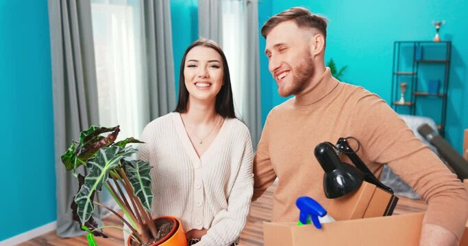 Portrait of young happy couple standing in the middle of new apartment. Handsome man holding box, pretty caucasian girl holding green flowerpot.