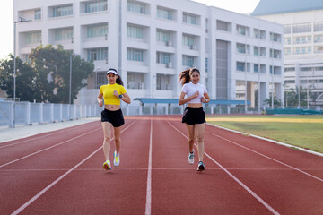 Fototapeta premium Two young Asian women in sports outfits jogging on running track in city stadium in the sunny morning to keep fitness and healthy lifestyle. Young fitness women run on the stadium track