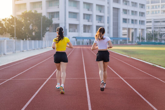 Two young Asian women in sports outfits jogging on running track in city stadium in the sunny morning to keep fitness and healthy lifestyle. Young fitness women run on the stadium track