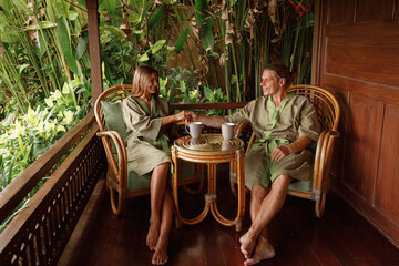 Couple in bathrobes enjoying  vacation, sit on terrace of wooden house, relax and drink tea