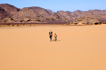 Fototapeta na wymiar Two Tourists And Their Camel Walking Across A Vast Sand Plain In The Sahara Desert, Libya