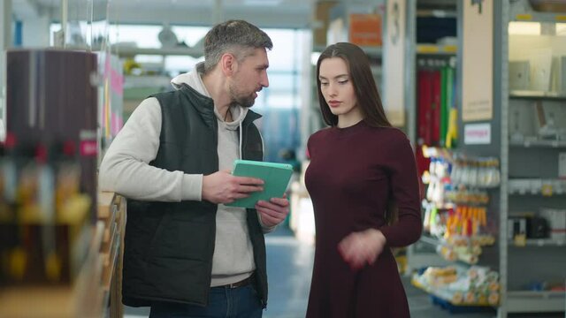 Confident Caucasian man consulting young charming woman choosing supplies in hardware store. Concentrated beautiful female shopper talking with male seller in building supplies shop.