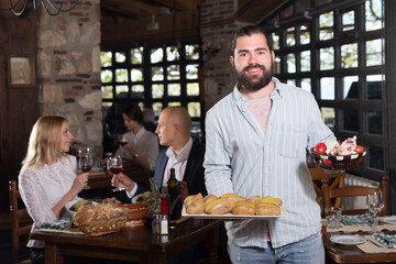 Fototapeta na wymiar Polite bearded waiter offering delicious dishes for tasting in cosy rustic restaurant ..