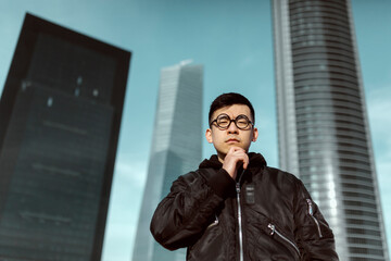 Chinese man, wearing round nerd glasses, with his hand on his face, with a thoughtful expression. With an skyscrapers in the background. Job, stock exchange, economy and chinese businessmen concept.