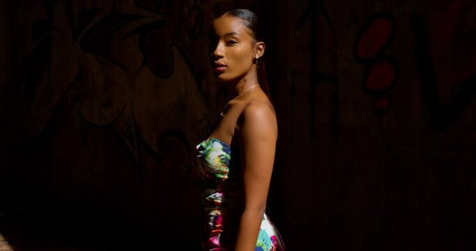 A young girl turns while standing in a ray of sunlight with grafitti on the wall in the bckground