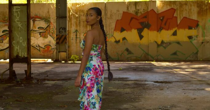Sexy Caribbean girl in a long flowing dress walks toward grafitti on the wall before turning around