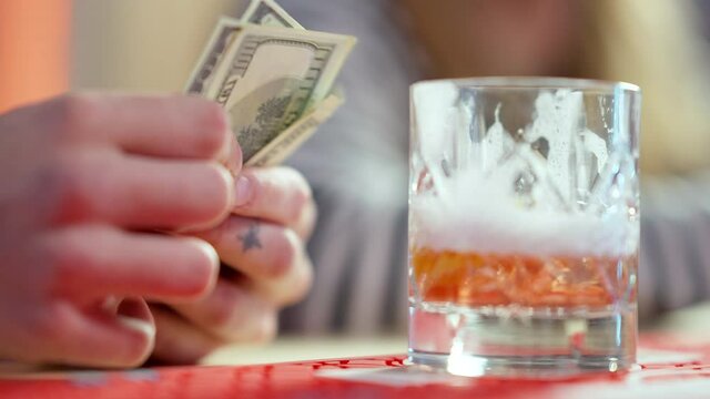 Glass with alcohol on bar counter with male hands counting dollars. Unrecognizable tattooed Caucasian man holding money and leaving. Payment for drinks.