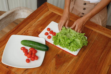Young trendy woman cooking healthy food at home kitchen - green salad from lettuce, chery tomato and cucumber, hands close up