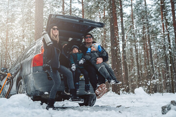 Vacations and enjoynment of a caucasian family. Three boys with their parents sit on car trunk in winter forest in daytime.