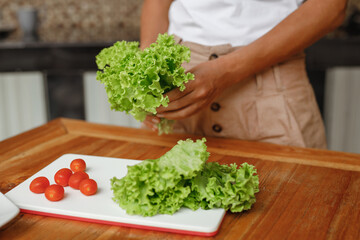 Young trendy woman cooking healthy food at home kitchen - green salad from lettuce, chery tomato and cucumber, hands close up