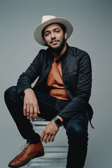 Manly indian hipster guy with beard and hat sits on chair posing in gray background and looking at camera.