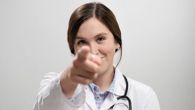 Doctor beckoning finger gesture. Happy positive smiling woman doctor on gray background looking at camera and points with finger and invite gesture asks to come to her. Slow motion