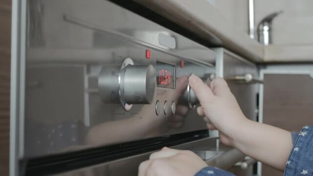 The little hands of a Caucasian teenage child turn on the timer by the oven to warm up the food. The child in the kitchen copes independently.