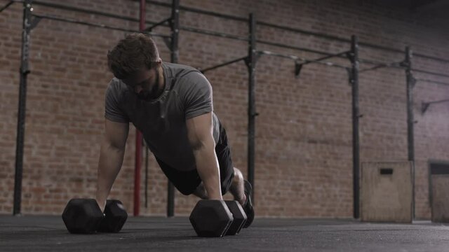 Wide shot of young Caucasian sportsman wearing sportswear doing push ups leaning on dumbbells in gym