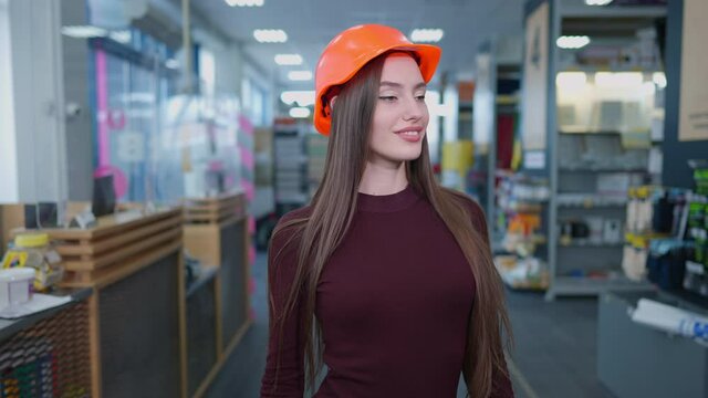 Portrait of elegant slim beautiful woman in hard hat posing in hardware store indoors. Charming brunette Caucasian buyer with brown eyes looking at camera smiling standing between rows with goods.