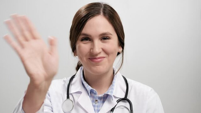 Doctor waving hello. Positive smiling woman doctor on gray background looking at camera and waving right hand showing greeting. Slow motion