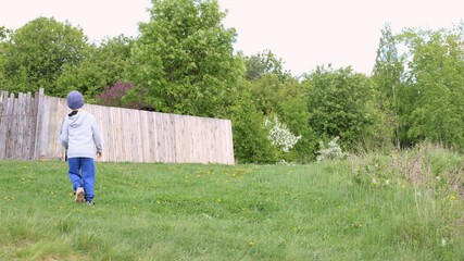 a boy walking in a green park or garden on a summer lawn in the countryside, a child walking alone in a green area along a village fence from the back, a lone kid walking on green grass