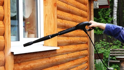 wet cleaning of the external wall of a residential private house from metal panels stylized as a log using a hand-held high pressure washer by a person in a blue uniform on a cloudy summer day