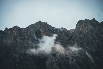 Atmospheric mountain landscape with low clouds on sharp rocks. Awesome rocky mountain with pointy top. Dark rocks in cloudy sky. Beautiful scenery with low clouds on rough crags. Pointed pinnacle.