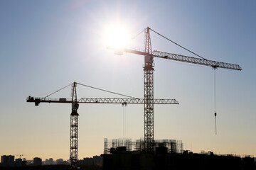 Construction cranes and unfinished residential building on blue sky and sunshine background. Housing construction, apartment block in city
