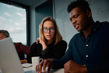 Caucasian and african female and male coworkers discussing something using laptop while sitting at...