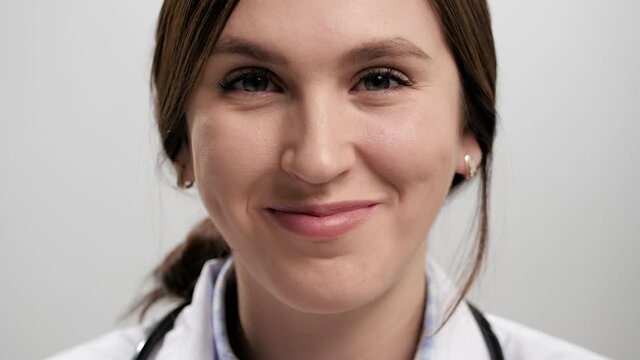 Doctor winks. Close-up of positive smiling woman doctor on gray background looking at camera and playfully winking with her right eye. Slow motion