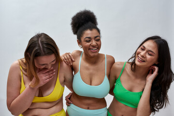 Three joyful diverse young women with different body shapes wearing colorful underwear laughing, posing together over light background