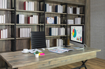 Workspace with desktop computer on wooden desk and black chair with bookshelf on the background.