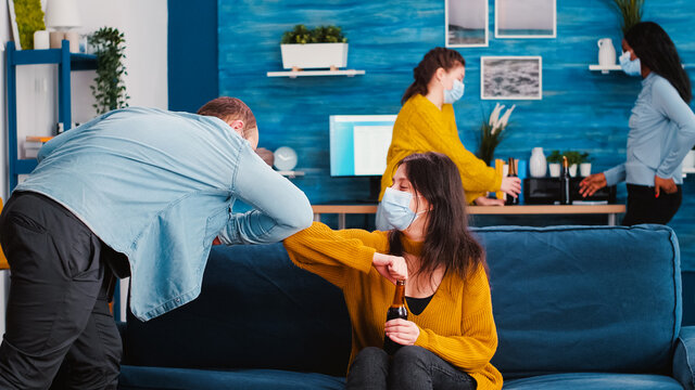 Friends In Protective Medical Face Mask Greet Their Elbows Keeping Social Distancing. New Greeting Elbow Bump Avoiding Spread Of Coronavirus Having Fun And Socializing At Home Party