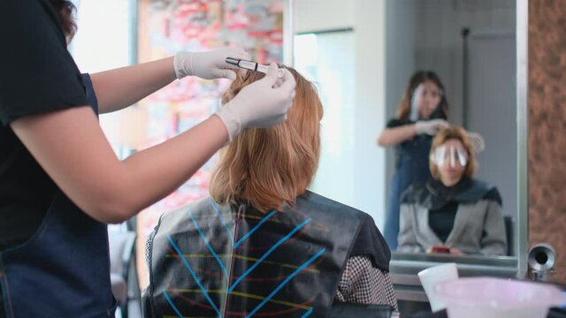 Asian Beauty salon barber woman prepare and start step for dye Caucasian customer hair in front of mirror and both of them wear face shield to protect virus infection with new normal lifestyle.