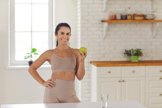 Active Healthy Lifestyle, Clean Eating Concept. Smiling Pretty Woman Athete In Sportswear Standing With Green Fresh Apple In Hands Before Or After Workout At Home