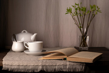A book, a vase with spring branches, white teapot and a cup on a table