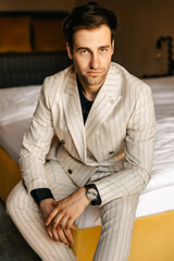 Fototapeta premium Handsome respectable groom is sitting on the bed of a luxury hotel room, the groom's light wedding suit