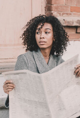 A woman of model African American appearance sits on a staircase and holds a newspaper in her hands against the background of a brick building