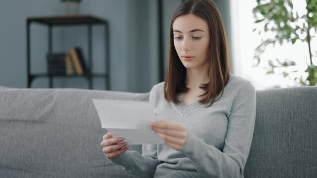 Charming young woman with brown hair sitting on grey couch and reading letter. Attractive female in casual outfit holding paper document in hands.