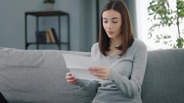 Excited young woman with brown hair reading paper document while sitting on couch at bright living room. Concept of correspondence and notification.