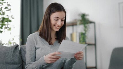 Cheerful young brunette in domestic clothes sitting on cozy sofa and reading letter. Concept of correspondence and good news.