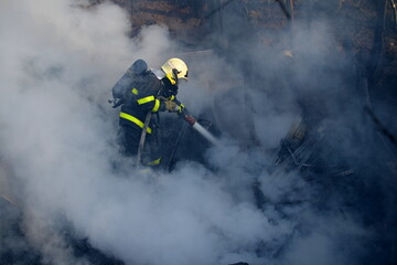 Firefighter with a breathing apparatus shrouded and surrounded by thick smoke extinguish the fire of wooden buildings with water from a hose