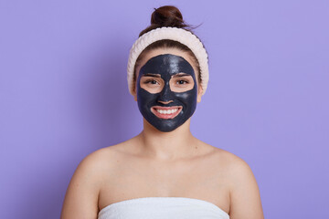 Close up portrait of beautiful woman with facial black mask isolated over lilac background, girl with white towel on her body and bare shoulders, satisfied and happy smile, doing cleansing procedure.