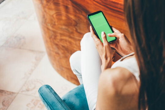 Back view unrecognizable lady types on smartphone at wooden bar counter in modern city restaurant. Close-up hand with manicure hold device with green screen sitting on high chair