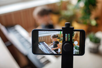 Pretty young musician in classic black dress playing digital piano at home during online concert and recording video, social distance during quarantine, online education concept, video blogging