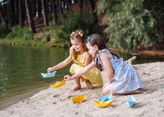 Two little girls launch origami paper boats into the river. The girls are dressed in blue and...