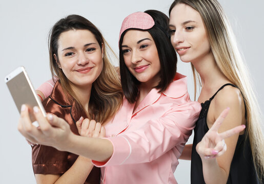 happy friends or teenage female wearing pajamas with smartphone taking selfie over grey background