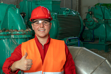 Young Worker in Red Coveralls and Hardhat Standing in Industrial Interior and Giving Thumbs Up